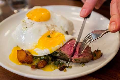 A piece of steak and two eggs on a plate and a fork and a knife cutting the steak