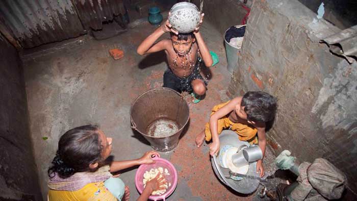 Child  bathing  with  a  bucket  of  water, another  child watching, and  mom  watching while preparing  food