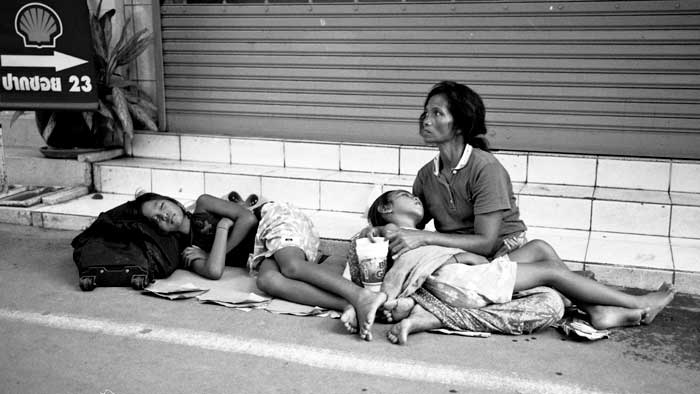 Mamá  sentada en la calle mientras sus 2 hijas duermen, la una en su regazo, la otra a su lado