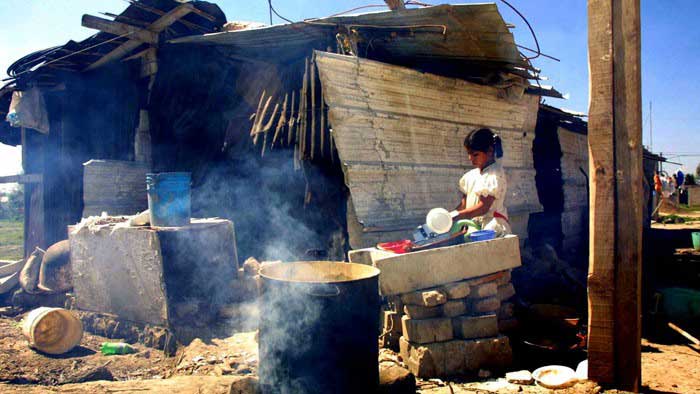 Niña lavando los platos afuera de una choza y humo saliendo del fuego con olla sobre él