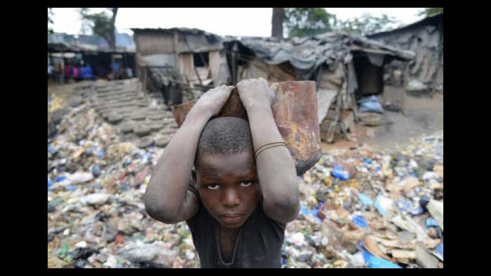 Niño caminando por un tugurio lleno de basura, cargando una basija de barro